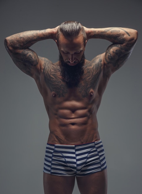 Foto gratuita hombre barbudo tatuado en bragas de rayas sosteniendo los brazos detrás de su cabeza. aislado sobre fondo gris.