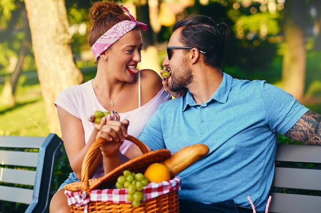 Hombre barbudo tatuado y mujer pelirroja están haciendo un picnic en un banco en un parque.