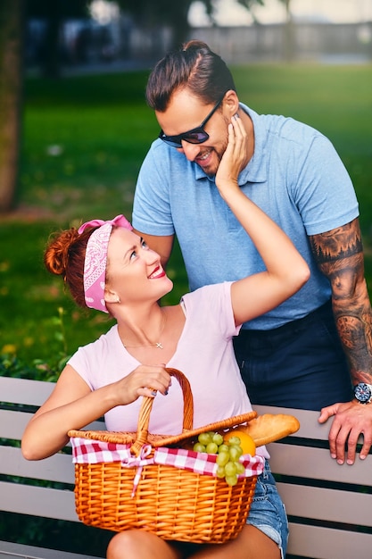Hombre barbudo tatuado y mujer pelirroja están haciendo un picnic en un banco en un parque.