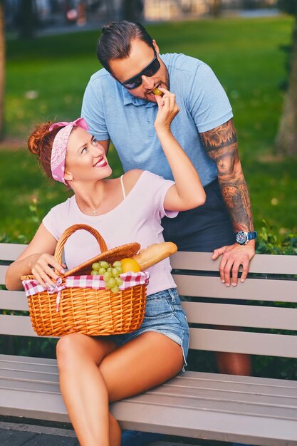 Hombre barbudo tatuado y mujer pelirroja están haciendo un picnic en un banco en un parque.