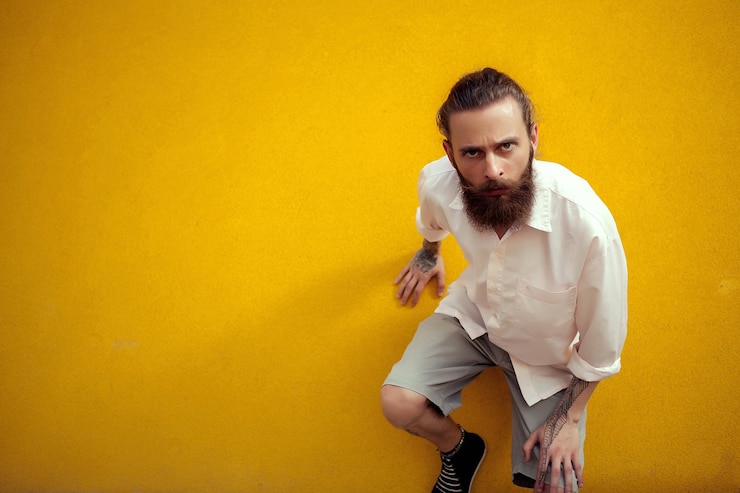 Foto gratuito hombre barbudo y tatuado posando al estilo de la moda en la pared amarilla exterior. moda y estilo