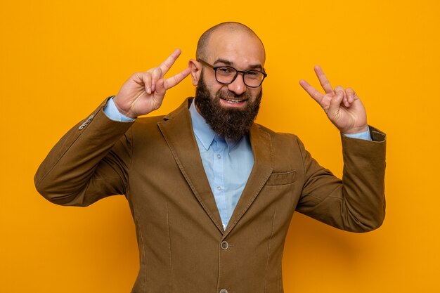 Hombre barbudo en traje marrón con gafas mirando a la cámara feliz y alegre sonriendo alegremente mostrando v-sign de pie sobre fondo naranja