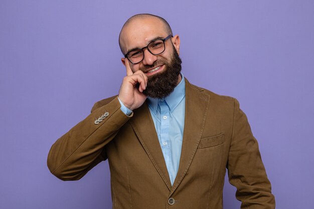 Hombre barbudo en traje marrón con gafas mirando a la cámara feliz y alegre sonriendo ampliamente de pie sobre fondo púrpura