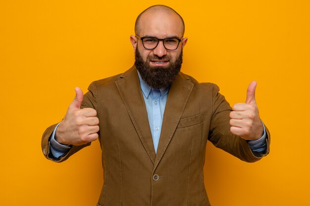 Hombre barbudo en traje marrón con gafas mirando feliz y alegre sonriendo ampliamente mostrando los pulgares para arriba