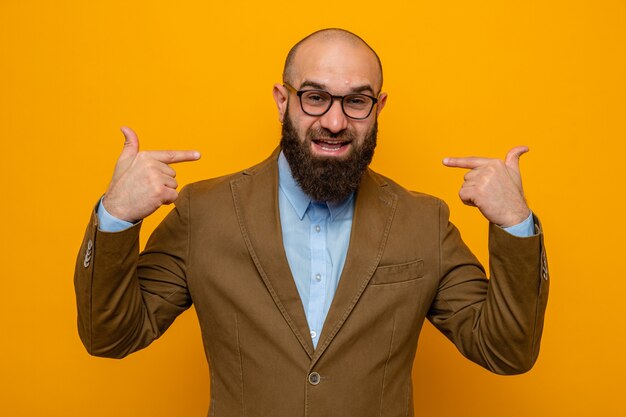 Hombre barbudo en traje marrón con gafas mirando feliz y positivo apuntando a sí mismo