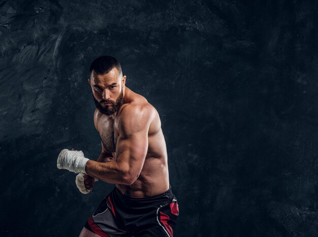 Hombre barbudo varonil con hermoso torso musculoso está posando para el fotógrafo en el fondo oscuro.