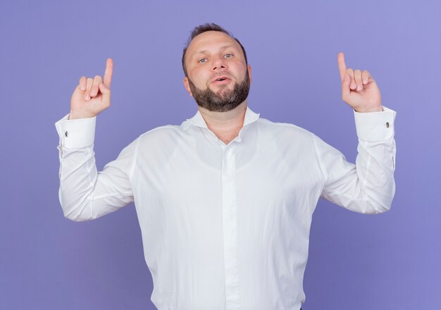 Hombre barbudo vestido con camisa blanca apuntando hacia arriba con los dedos índices mirando con cara seria de pie sobre la pared azul