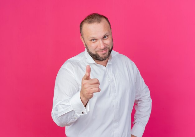 Hombre barbudo vestido con camisa blanca apuntando con el dedo índice sonriendo confiado de pie sobre la pared rosa