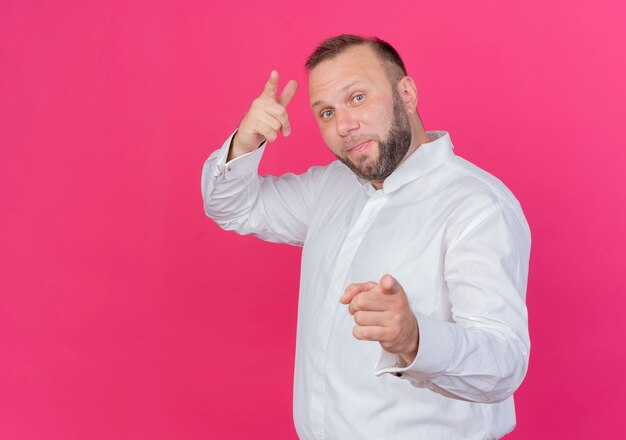 Hombre barbudo vestido con camisa blanca apuntando con los dedos índices de pie sobre la pared rosa