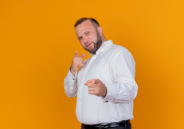 Hombre barbudo vestido con camisa blanca apuntando con los dedos sonriendo alegremente de pie sobre la pared naranja