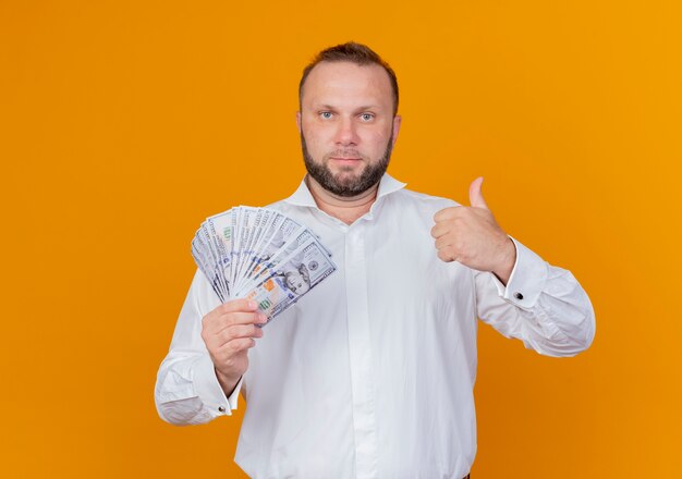 Hombre barbudo vestido con camisa blanca con dinero en efectivo mostrando los pulgares para arriba sobre la pared naranja