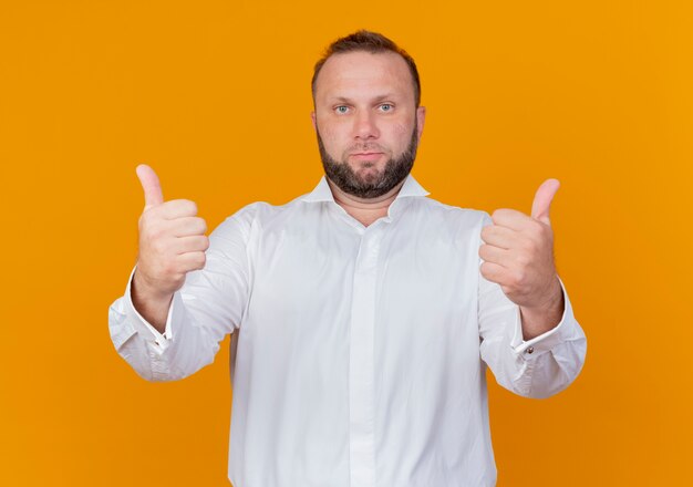 Hombre barbudo vestido con camisa blanca con expresión de confianza mostrando los pulgares para arriba de pie sobre la pared naranja