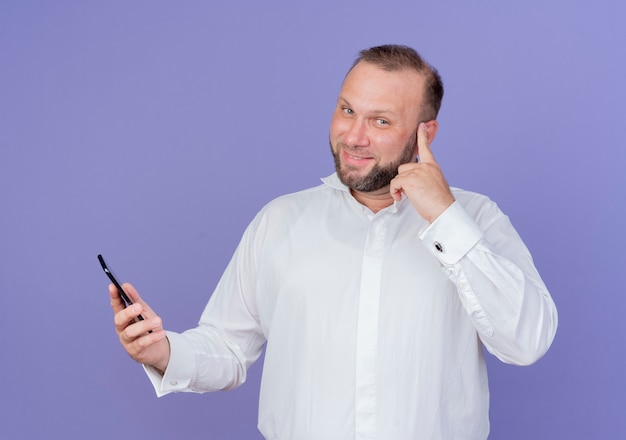 Hombre barbudo vestido con camisa blanca hablando por teléfono móvil mirando sonriendo señalando templo centrado en una tarea de pie sobre la pared azul