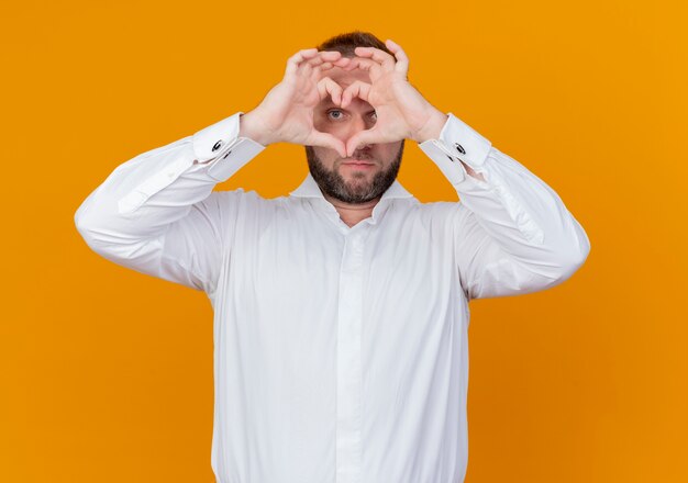 Hombre barbudo vestido con camisa blanca haciendo gesto de corazón con los dedos mirando a través de los dedos de pie sobre la pared naranja