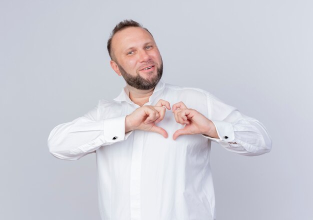 Hombre barbudo vestido con camisa blanca haciendo gesto de corazón con los dedos sonriendo de pie sobre la pared blanca