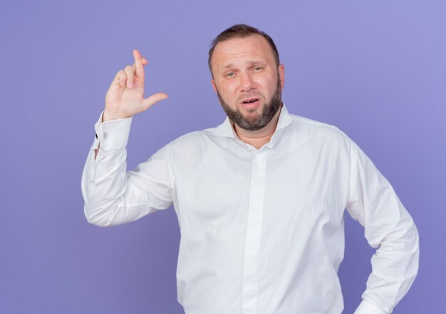 Hombre barbudo vestido con camisa blanca haciendo una promesa cruzando los dedos de pie sobre la pared azul