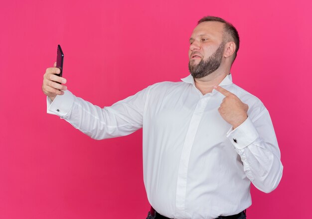Hombre barbudo vestido con camisa blanca haciendo selfie apuntando con el dedo sobre la pared rosa