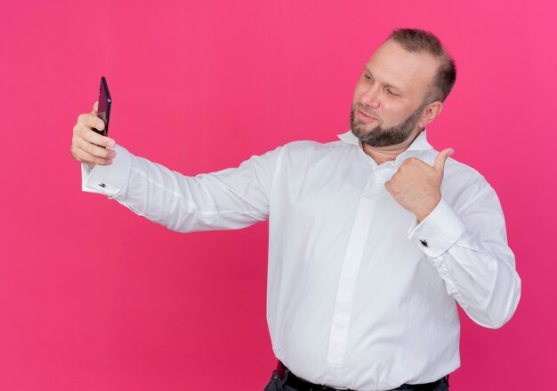 Hombre barbudo vestido con camisa blanca haciendo selfie mostrando los pulgares para arriba de pie sobre la pared rosa