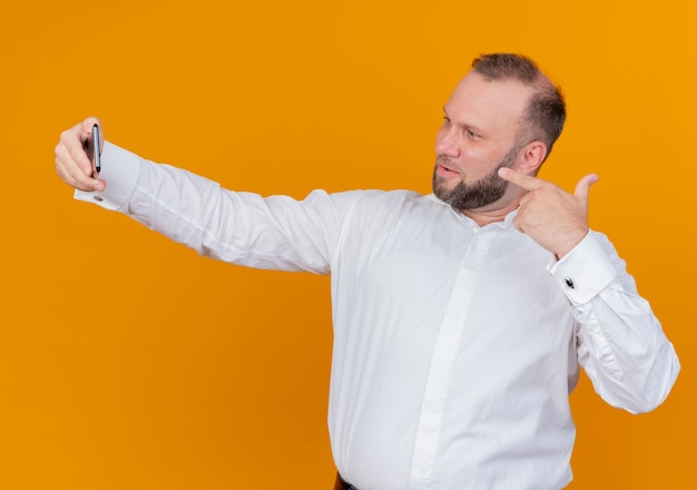 Hombre barbudo vestido con camisa blanca haciendo selfie sonriendo de pie sobre la pared naranja