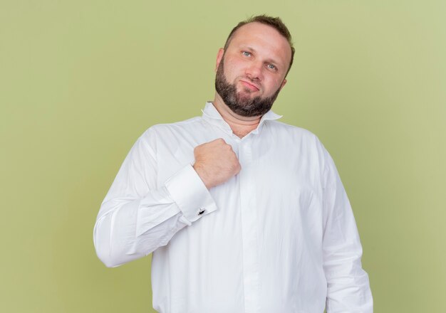 Hombre barbudo vestido con camisa blanca mirando confiado con finst en su pecho parado sobre pared ligera