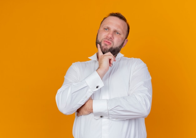 Hombre barbudo vestido con camisa blanca mirando a un lado desconcertado parado sobre la pared naranja