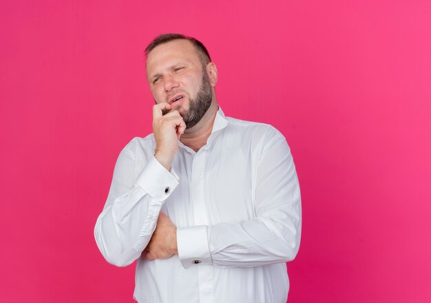 Hombre barbudo vestido con camisa blanca mirando a un lado perplejo parado sobre pared rosa