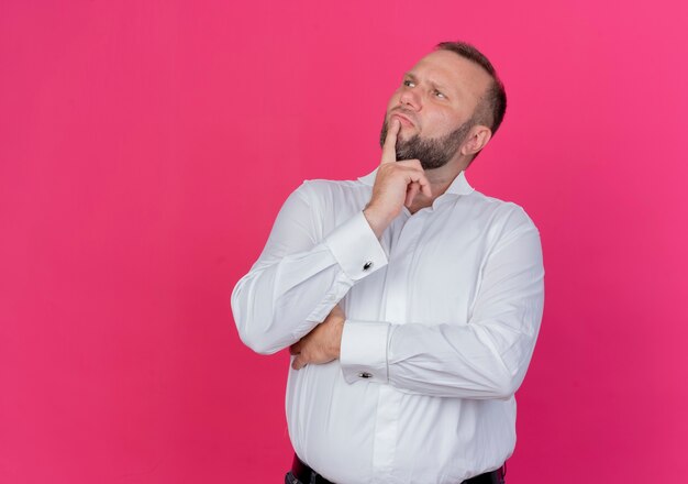 Hombre barbudo vestido con camisa blanca mirando a un lado perplejo parado sobre pared rosa