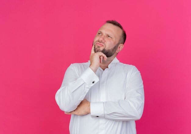 Hombre barbudo vestido con camisa blanca mirando a un lado perplejo parado sobre pared rosa