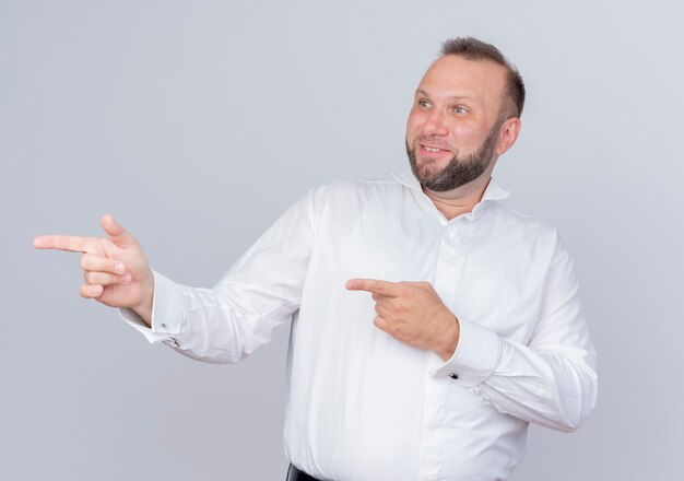 Hombre barbudo vestido con camisa blanca mirando a un lado sonriendo apuntando con el dedo índice hacia el lado de pie sobre la pared blanca