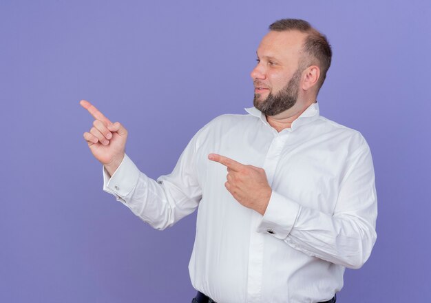 Hombre barbudo vestido con camisa blanca mirando a un lado con una sonrisa pointign con los dedos índices hacia el lado de pie sobre la pared azul