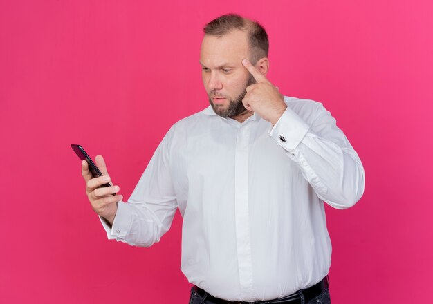 Hombre barbudo vestido con camisa blanca mirando la pantalla de su teléfono inteligente apuntando su sien se centró en una tarea difícil de pie sobre la pared rosa