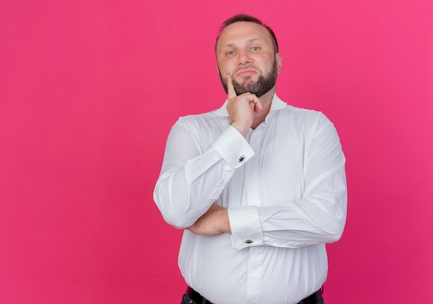 Hombre barbudo vestido con camisa blanca mirando perplejo parado sobre pared rosa