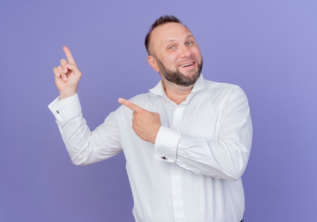 Hombre barbudo vestido con camisa blanca mirando sonriendo con cara feliz apuntando con los dedos hacia el lado de pie sobre la pared azul