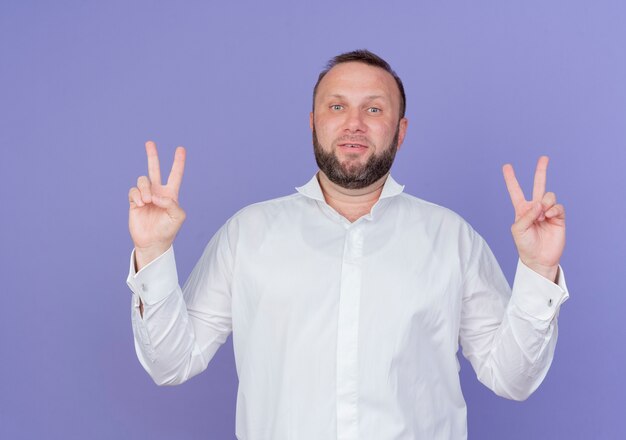 Hombre barbudo vestido con camisa blanca mirando sonriendo mostrando v-sign de pie sobre la pared azul