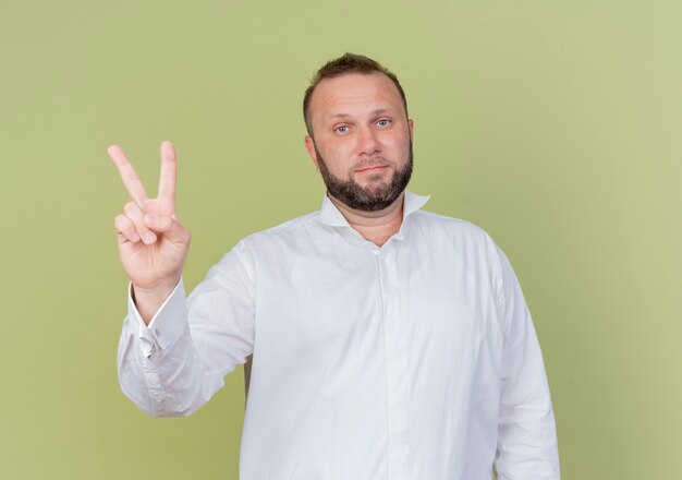 Hombre barbudo vestido con camisa blanca mostrando y apuntando hacia arriba con los dedos número dos mirando con cara seria de pie sobre una pared de luz