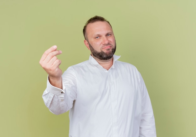 Hombre barbudo vestido con camisa blanca mostrando gesto de dinero frotando los dedos de pie sobre la pared de luz