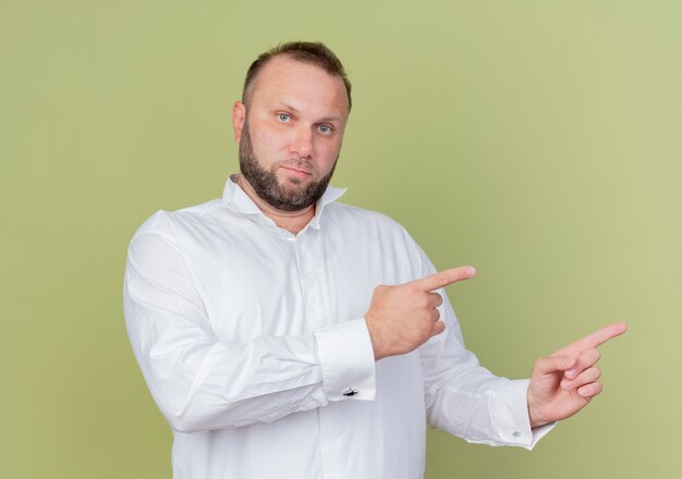 Hombre barbudo vestido con camisa blanca con rostro serio apuntando con los dedos índices hacia el lado de pie sobre la pared de luz