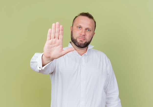 Hombre barbudo vestido con camisa blanca con rostro serio haciendo señal de stop con la mano abierta de pie sobre la pared de luz