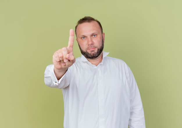 Hombre barbudo vestido con camisa blanca con rostro serio que muestra la advertencia del dedo índice de pie sobre la pared de luz