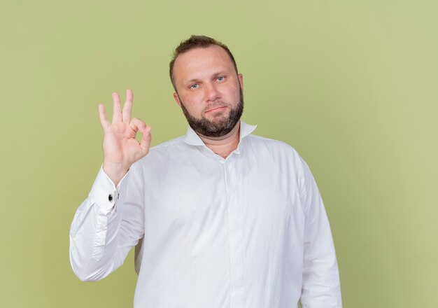 Hombre barbudo vestido con camisa blanca sonriendo confiado mostrando signo ok de pie sobre la pared ligera