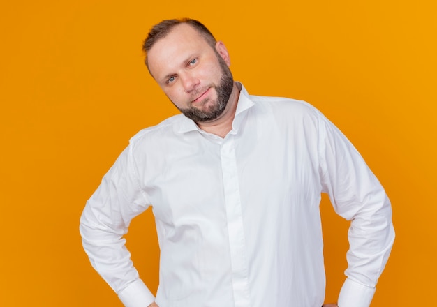 Hombre barbudo vestido con camisa blanca sonriendo confiado de pie sobre la pared naranja