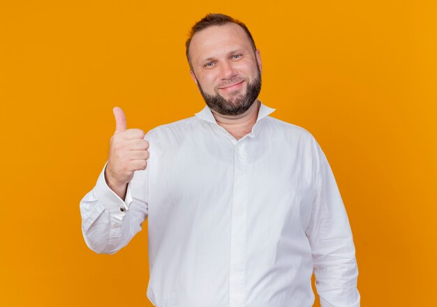 Hombre barbudo vestido con camisa blanca sonriendo mostrando los pulgares para arriba de pie sobre la pared naranja