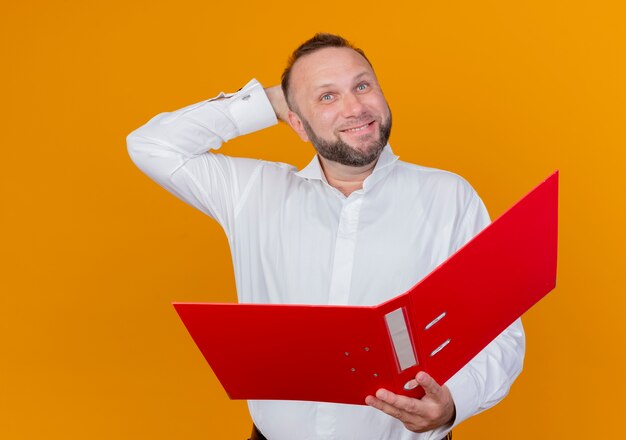 Hombre barbudo vestido con camisa blanca sosteniendo la carpeta mirando a un lado con una sonrisa en la cara de pie sobre la pared naranja