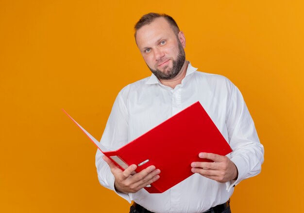 Hombre barbudo vestido con camisa blanca sosteniendo una carpeta sonriendo de pie sobre la pared naranja