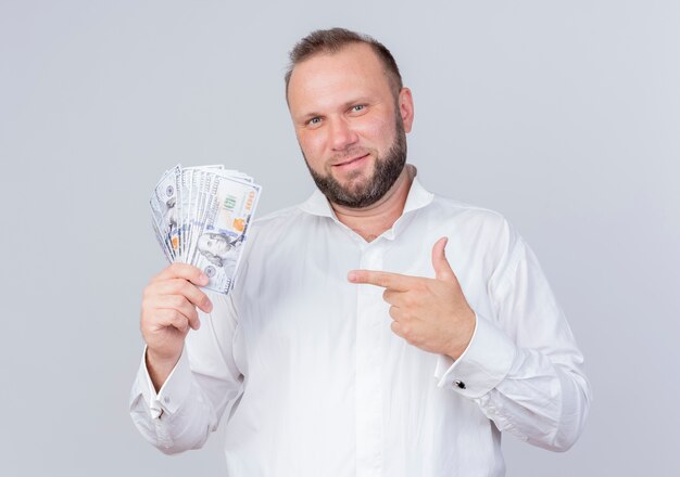 Hombre barbudo vestido con camisa blanca sosteniendo dinero en efectivo apuntando con el dedo índice al dinero sonriendo confiado de pie sobre la pared blanca