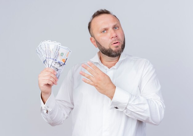 Hombre barbudo vestido con camisa blanca sosteniendo dinero en efectivo mostrando y apuntando hacia arriba con los dedos número cuatro mirando sorprendido de pie sobre la pared blanca