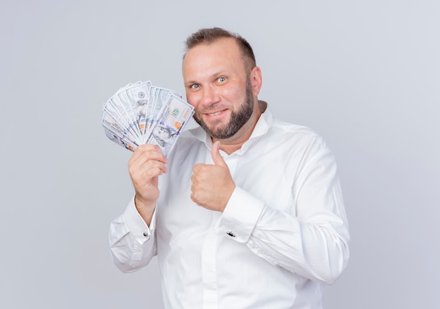 Hombre barbudo vestido con camisa blanca sosteniendo dinero en efectivo sonriendo astutamente mostrando los pulgares para arriba sobre la pared blanca