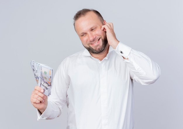 Hombre barbudo vestido con camisa blanca sosteniendo dinero en efectivo sonriendo con cara feliz de pie sobre la pared blanca