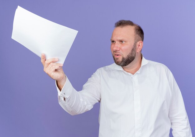 Hombre barbudo vestido con camisa blanca sosteniendo una hoja de papel en blanco mirando a un lado disgustado de pie sobre la pared azul