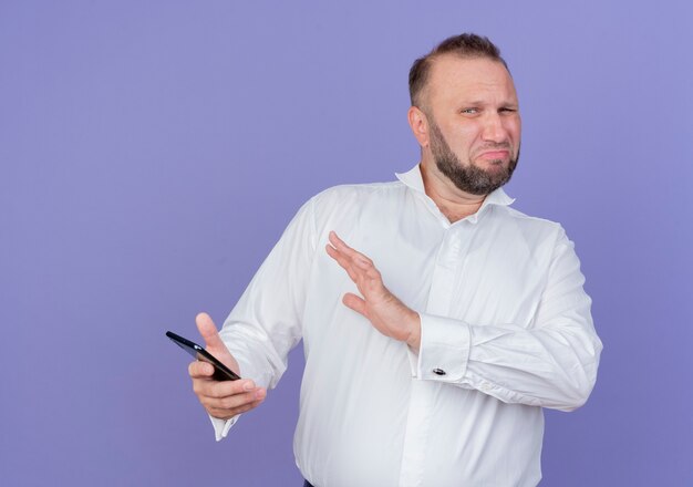 Hombre barbudo vestido con camisa blanca sosteniendo smartphone haciendo gesto de defensa con la mano mirando con expresión de disgusto de pie sobre la pared azul